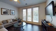 This single-floor residence offers a thoughtfully designed layout that balances open communal areas with comfortable private spaces. The living room serves as a welcoming focal point, featuring dark hardwood-style flooring, a ceiling fan, and a striking set of full-height glass doors that flood the space with natural light and provide outdoor views of surrounding greenery and neighboring buildings. Horizontal blinds offer privacy as needed.

The kitchen is well-equipped with warm honey-toned cabinetry, stainless steel appliances including a refrigerator, range, and microwave, and a granite-topped island with seating for two. Pendant lighting above the island adds a refined touch, while the open sightlines to the living and dining areas create an inviting, connected flow.

The bedroom is carpeted for comfort and includes a ceiling fan, a window with natural light, and ample space for a full furniture arrangement. A patterned accent chair adds character to the room.

The bathroom features rich dark wood-look tile flooring, warm-toned cabinetry with a light countertop, a large mirror, and overhead cabinetry for storage. The overall aesthetic of the space is cohesive and contemporary, with neutral tones creating a calm, livable atmosphere throughout.