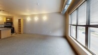 Located in the heart of Des Moines at 333 E Grand Avenue, Unit 205 is a thoughtfully laid-out single-floor residence with a combined kitchen and living area, plus one full bathroom. The open-plan living and kitchen space features a bold black-and-white checkered tile floor in the kitchen zone that transitions to plush wall-to-wall carpet in the living area. The kitchen is equipped with black appliances — including a full-size refrigerator, range, microwave, and dishwasher — complemented by warm wood cabinetry, a dark countertop island with pendant lighting overhead, and a built-in open shelving unit. Exposed industrial-style ductwork runs along the ceiling, lending the space an urban loft character. Large multi-pane windows along the living area walls allow abundant natural light and offer views of the surrounding cityscape, enhancing the open feel of the space. An accent wall of rich orange-toned wood paneling adds a striking visual focal point. The bathroom is clean and functional, featuring a full soaking tub and shower combination, wood-look tile flooring, and neutral wall tones. Wall-mounted sconce lighting throughout adds a warm, ambient quality to each area of this efficiently designed urban residence.