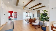 Located at 4896 Stellar Drive in St. Cloud, Florida, this single-floor amenity space combines a sophisticated lounge area with a dedicated fitness center, connected by a transitional hallway. The lounge area makes an immediate impression with its elevated ceilings adorned with dark wood exposed beams and dramatic sputnik-style gold chandeliers that cast warm, ambient light throughout. Light hardwood flooring flows seamlessly underfoot, while sheer white curtains filter abundant natural light from large windows and glass entry doors. A bold textured accent wall featuring a circular relief pattern anchors the lounge's media wall, complementing the mid-century modern furnishings that include a plush sectional sofa, sculptural gold coffee tables, and a geometric area rug. French glass doors lead directly into the fitness center, which offers a striking contrast with its bold red accent wall and a large-scale motivational typographic mural on a wood-paneled backdrop. The fitness room is equipped with a variety of cardio and strength training machines, including ellipticals, a stair climber, and weight equipment, all set on durable rubberized flooring. Natural light enters through windows overlooking the outdoor surroundings. Together, these spaces deliver a refined yet functional environment ideal for community gathering and wellness.