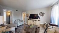 This single-story residence is thoughtfully laid out across a compact yet functional floor plan. The living room serves as the heart of the home, featuring warm neutral-painted walls, durable wood-look flooring, and ample natural light filtering through windows dressed with sheer curtains. A light pine media console anchors the entertainment area, complemented by a rustic wooden coffee table and comfortable seating. A decorative gear wall accent adds an industrial touch to the space.

The galley-style kitchen is efficiently designed with white cabinetry, dark countertops, a double sink, and white appliances including a gas range and refrigerator. A patterned area rug adds visual interest underfoot. Adjacent to the kitchen, a dining nook offers space for a round table and chairs, accented by a multi-panel black-and-white nature-themed wall art display and a window providing natural light.

The bedroom is a calm retreat with a closet, natural window light, wood-look flooring, and neutral tones. An additional flex room with a large bifold closet provides versatile living options. The bathroom features a tub-shower combination, white cabinetry, dark countertops, and subway tile detailing, completing this well-appointed home.