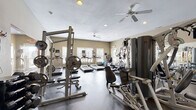This single-floor fitness center is thoughtfully designed to accommodate a full range of workout routines. The space is divided into distinct functional zones, seamlessly flowing from cardio to strength training areas. Along one windowed wall, a row of treadmills and elliptical machines are positioned to take advantage of natural light streaming in through multiple large windows, each equipped with individual monitor screens. Ceiling fans are installed throughout, providing consistent air circulation during workouts.

The strength training section features a power rack with an extensive plate weight collection, a multi-function cable machine, and an adjustable bench, offering versatility for various resistance exercises. A tiered dumbbell rack holds a complete set of free weights, complemented by a dedicated stretching and accessory area. Full-length mirrors line one wall, visually expanding the space and allowing users to monitor their form during exercise.

The flooring transitions between rubberized mats in the weight area and a smooth, durable surface in the cardio zone. Recessed lighting supplements natural light throughout, while a wall-mounted flat-screen television adds an entertainment option. A restroom is conveniently accessible directly from the fitness floor. The overall ambiance is practical and well-organized, designed to support a comprehensive fitness experience.