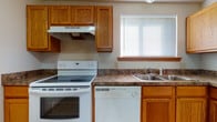 Located at 5371 Big Tyler Rd in Cross Lanes, West Virginia, this single-floor unit offers a straightforward and functional layout well-suited for comfortable everyday living. The kitchen is equipped with warm oak cabinetry providing ample storage, dark laminate countertops, and white appliances including an electric range with overhead ventilation, a dishwasher, and a double-basin stainless steel sink positioned beneath a window that allows natural light to brighten the space. The tile flooring in the kitchen is easy to maintain and transitions into the open living area, which features neutral wall-to-wall carpeting and a wide entryway connecting the two spaces. The living area is generously proportioned with clean, neutral-toned walls and a simple, open feel. The bedrooms are carpeted and each features a window with blinds, offering natural light and a quiet atmosphere. The bathroom includes a full tub-and-shower combination with chrome fixtures, a toilet, and tile flooring in a warm terracotta tone, finished with wood-trimmed doorways that echo the oak accents found throughout the unit. Overall, the space presents a clean, neutral palette with consistent warm wood detailing throughout.