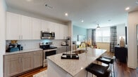 This single-floor residence is designed with a contemporary open-concept layout that seamlessly connects the kitchen, dining, and living areas. The kitchen is a standout feature, equipped with stainless steel appliances including a French door refrigerator, electric range, and built-in microwave. Two-tone cabinetry pairs crisp white upper cabinets with warm taupe lower cabinets, complemented by a classic white subway tile backsplash and light granite countertops. A generously sized island with an undermount sink and bar seating serves as both a functional workspace and a social hub.

Rich hardwood-style flooring flows throughout the entire unit, adding warmth and continuity. The living area benefits from multiple windows fitted with blinds and patterned drape panels, allowing ample natural light while maintaining privacy. A ceiling fan keeps the space comfortable year-round.

The bedroom is a calm retreat, featuring soft blue-grey walls, large windows, and continued hardwood-style flooring. The bathroom offers granite countertops, wood-toned cabinetry, a tub-shower combination, and direct access to a spacious walk-in closet with built-in shelving. Recessed lighting throughout enhances the modern, polished ambiance of the entire unit.