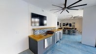 This single-floor space is designed with entertainment and gathering in mind, blending modern functionality with a relaxed coastal aesthetic. The wet bar area serves as a central focal point, outfitted with warm wood-toned countertops, a built-in sink, stainless steel under-counter refrigerators, and a blue and white mosaic tile backsplash that adds a nautical touch. A large wall-mounted flat-screen TV is positioned directly above the bar, making it an ideal setup for watching events or presentations while guests gather.

The flooring throughout is polished concrete in a cool blue-grey tone, contributing to the contemporary, open feel of the space. Oversized ceiling fans — including a striking multi-blade fan overhead — ensure comfortable airflow throughout. The open-plan layout transitions seamlessly into a lounge and dining area furnished with casual seating and dining tables, illuminated by recessed lighting. Coastal-themed artwork decorates the walls, reinforcing the relaxed atmosphere. The overall design balances practicality with style, making this a versatile venue well-suited for social gatherings, events, or communal use.
