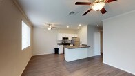 This single-floor space offers a clean, modern aesthetic defined by an open-concept layout that seamlessly connects the kitchen and living area. The kitchen is a standout feature, outfitted with white shaker-style cabinetry, dark countertops, and a full suite of stainless steel appliances including a refrigerator, range, and microwave. A peninsula with an integrated sink extends into the open living space, providing additional prep space and a natural gathering point. The flooring throughout is rich, dark hardwood-style planking that grounds the space and adds warmth against the light, neutral-toned walls. Recessed lighting in the kitchen is complemented by ceiling fans in both the kitchen and living areas, ensuring comfortable airflow and well-distributed light. Crown molding along the upper walls adds a refined architectural touch, while large windows allow natural light to filter in. The overall ambiance is fresh and move-in ready, combining practical design with contemporary style in a cohesive, comfortable single-level layout.