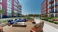 This expansive outdoor courtyard serves as a central amenity space nestled between multi-story residential buildings featuring distinctive red and gray siding with white-trimmed windows. The space is thoughtfully organized into several functional zones, each offering a unique outdoor experience.

A generous lounge area anchors one section of the courtyard, furnished with a large blue sectional sofa and a sleek fire pit table set atop wood-look porcelain tile decking. Adjacent to this is a shaded pergola zone equipped with a retractable canopy, beneath which oversized blue daybed-style loungers provide a comfortable retreat. Cobalt accent stools and ornamental grasses in raised planters add vibrant color and texture throughout.

The outdoor kitchen area features a built-in grill with granite countertops and bar-height seating, ideal for al fresco dining. Raised planter beds filled with native grasses, flowering shrubs, and ornamental trees are integrated throughout the space, softening the hardscape and adding a naturalistic quality. A dedicated pool area with lounge seating is visible toward the rear of the courtyard.

Large-format stone pavers and warm wood-tone decking unify the various zones, while the open-air design allows abundant natural light to permeate the space throughout the day, creating a bright and welcoming outdoor environment.