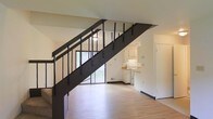 This two-story residence is designed with a straightforward, functional layout that makes efficient use of its space across both levels. The main floor immediately draws attention with its light-toned hardwood flooring, which lends warmth and continuity to the open living area. A striking dark metal staircase serves as a central architectural feature, its industrial-style vertical balusters and solid stringer creating a strong visual contrast against the neutral white walls and pale floors. The open riser design of the staircase allows light to pass through, contributing to an airy feel on the main level. A large sliding glass door at the rear of the main floor floods the space with natural light and provides a view of the greenery outside, further enhancing the open atmosphere. The kitchen, visible from the living area, features white cabinetry, a light-toned countertop, and white appliances, offering a clean and practical cooking space. The walls throughout are finished in a neutral off-white tone, providing a versatile backdrop. The second floor, accessible via the metal staircase, extends the living space further. Overall, the residence presents a no-frills, move-in-ready environment with a blend of industrial and casual design elements.