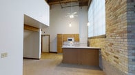 This two-story space showcases an impressive industrial loft aesthetic, defined by dramatically high ceilings that draw the eye upward and create an expansive sense of volume throughout the main living area. The open-concept kitchen serves as a central feature, equipped with a peninsula counter, oak-toned upper cabinetry, a white appliance suite including a range and refrigerator, and pendant lighting suspended from above. A ceiling fan adds both function and visual interest at height. Exposed brick along one wall introduces a raw, textural element that complements the space's industrial character, while large translucent windows along the same wall diffuse natural light softly into the interior. The flooring transitions between carpet in the living area and hardwood-style flooring near the entry corridor, adding warmth and variety underfoot. The two-story layout accommodates four bedrooms and two bathrooms, with the upper level accessible from the main floor. The overall ambiance balances utilitarian, loft-inspired architecture with functional residential amenities, making it a distinctive and characterful space suited for multi-occupant living.