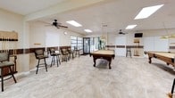 This single-floor recreation space is designed for social gatherings and leisure activities, offering a well-appointed and open layout throughout. The centerpiece of the room is a classic wood-framed billiards table, illuminated by a hanging pendant light that draws the eye and anchors the space. Large windows along one wall flood the interior with natural light, providing views of lush outdoor greenery and creating an airy, inviting atmosphere. The flooring throughout is a light-toned tile with a subtle stone pattern, contributing to the room's clean and polished aesthetic. A decorative wainscoting border in a warm brown tone runs along the lower portion of the walls, adding a refined architectural detail. Ceiling fans are strategically placed to ensure comfortable airflow across the space. Bar-height swivel chairs with cushioned seats and ornate wrought-iron frames are arranged along the walls, offering ample seating for spectators and socializers alike. A dedicated dart area is tucked into one corner, complete with a mounted dartboard, cork scoreboard panels, and additional bar seating. A bulletin board near the entrance provides a practical information hub. Recessed panel ceiling lights supplement the natural light, ensuring the space remains bright and functional throughout the day.