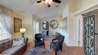 This 2-bedroom, 2-bathroom single-floor residence offers a well-appointed layout designed for comfortable everyday living. The open-concept living area features warm neutral tones, plush carpeting, and vaulted ceilings accented by a ceiling fan with integrated lighting, creating an airy and inviting atmosphere. Large windows dressed with patterned curtains allow generous natural light to fill the space, enhancing the bright and welcoming ambiance. The living room is styled with ample space for seating arrangements and connects seamlessly to the dining and kitchen area. A breakfast bar with bar stools provides a casual dining option, while a pass-through opening maintains visual connectivity between the kitchen and the main living space. Decorative elements, including an oversized round wall clock and framed artwork, contribute a contemporary aesthetic throughout. The kitchen features dark cabinetry and pendant lighting, adding a sleek contrast to the lighter walls. With two well-sized bedrooms and two full bathrooms, this residence provides both privacy and functionality. The thoughtful floor plan, combined with stylish finishes and an open flow between living spaces, makes this home an attractive and practical option for a variety of lifestyles.