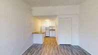 This single-story unit offers a clean, functional layout designed to maximize the available living space. The main living area is bright and open, featuring wood-look laminate flooring that runs throughout the entire unit, white walls, and a textured ceiling that adds subtle character. A large window in the living area allows natural light to fill the room, while a wall-mounted HVAC unit provides climate control.

The kitchen is thoughtfully arranged and separated from the living space by a small breakfast bar with a laminate countertop — a convenient feature for casual dining or extra prep space. White shaker-style cabinetry extends to the ceiling, offering generous storage. The kitchen is equipped with a white refrigerator, an electric range, a range hood, and a single-basin sink with stone-look countertops below white lower cabinets.

The bedroom is a well-proportioned, light-filled room with clean white walls and a set of louvered double closet doors providing ample storage. The bathroom is neatly appointed with a single vanity featuring a white sink basin, a mirror, a toilet, and grab bars installed along the wall for added accessibility. The unit's straightforward, move-in-ready design makes it a practical and comfortable living space.