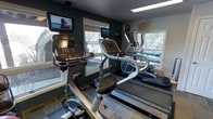 This fitness center is designed to provide a focused and motivating workout environment. The space is anchored by a suite of modern cardio equipment, including upright bikes, elliptical trainers, and Star Trac treadmills, each outfitted with individual flat-screen entertainment monitors for an engaging exercise experience. The room is finished with dark, richly toned walls that create a dynamic and energetic atmosphere, complemented by clean tile flooring throughout. Natural light fills the space generously through multiple large windows positioned along the walls, offering pleasant views of a wooded, outdoor landscape that enhances the sense of openness. A flush ceiling light panel provides consistent overhead illumination, ensuring the space remains bright and functional at any time of day. The windows also provide a visual connection to an outdoor patio or terrace area, visible through the glass, further adding to the welcoming ambiance. The layout is thoughtfully arranged to allow comfortable movement between machines while maximizing the use of available floor space. Overall, this fitness center balances practical functionality with an inviting aesthetic, making it a well-rounded facility for cardiovascular training.