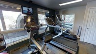 This fitness center is designed to provide a focused and motivating workout environment. The space is anchored by a suite of modern cardio equipment, including upright bikes, elliptical trainers, and Star Trac treadmills, each outfitted with individual flat-screen entertainment monitors for an engaging exercise experience. The room is finished with dark, richly toned walls that create a dynamic and energetic atmosphere, complemented by clean tile flooring throughout. Natural light fills the space generously through multiple large windows positioned along the walls, offering pleasant views of a wooded, outdoor landscape that enhances the sense of openness. A flush ceiling light panel provides consistent overhead illumination, ensuring the space remains bright and functional at any time of day. The windows also provide a visual connection to an outdoor patio or terrace area, visible through the glass, further adding to the welcoming ambiance. The layout is thoughtfully arranged to allow comfortable movement between machines while maximizing the use of available floor space. Overall, this fitness center balances practical functionality with an inviting aesthetic, making it a well-rounded facility for cardiovascular training.