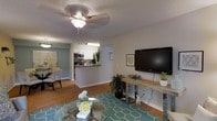 This single-floor residence offers a comfortable and functional layout designed for everyday living. The open-concept living area serves as the heart of the home, featuring warm hardwood flooring, a ceiling fan with light fixture, and a textured ceiling that adds subtle character. A wall-mounted flat-screen TV and a console table anchor the living space, while a glass coffee table and a vibrant teal geometric area rug bring color and style to the room.

The living area flows naturally into the kitchen, which is separated by a convenient breakfast bar with dark countertops — ideal for casual dining or entertaining. The kitchen is equipped with modern stainless steel appliances, including a microwave and oven range, and is complemented by crisp white cabinetry that maximizes storage. A decorative plant adds a fresh, natural touch to the kitchen counter.

Adjacent to the living space, a dining area accommodates a full dining set, maintaining the open and connected feel of the floor plan. Throughout the home, neutral wall tones create a calm, inviting ambiance, while the hardwood floors unify each area seamlessly. This well-appointed space balances practicality with a welcoming aesthetic.