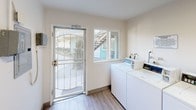 This shared laundry facility is a single-floor space designed for practical, everyday use. The room is finished with light wood-look flooring and bright white walls that create a clean, airy atmosphere. Natural light enters through a sliding window and a security-screened exterior door, which also provides direct access to the outdoor common area — visible through the door's glass panels, offering views of a courtyard and stairwell.

The laundry area is equipped with multiple top-loading washing machines and a combination of stacked washer-dryer units, accommodating a high volume of laundry needs. A standout feature is the generous granite-topped folding table that runs along one wall, providing ample surface area for sorting and folding laundry. Exposed plumbing connections and utility hookups are neatly integrated into the space.

Safety is addressed with a wall-mounted fire extinguisher station. The ceiling features exposed conduit, adding a utilitarian character consistent with the room's functional purpose. The overall layout is open and well-organized, allowing for easy movement between machines and the folding station. The combination of durable, low-maintenance finishes and practical amenities makes this a functional and well-appointed shared laundry room.