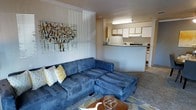 This single-floor residential space offers a practical and welcoming layout designed for comfortable everyday living. The living area serves as the heart of the home, featuring plush, oversized sectional seating and a decorative area rug that adds warmth and texture underfoot. Wall art featuring an abstract nature-inspired composition adds a touch of personality and color to the space. A partial wall with vertical paneling detail provides a subtle architectural divider while maintaining an open, connected feel throughout.

The kitchen is accessible directly from the living area, separated by a countertop breakfast bar that doubles as a casual dining surface. White upper cabinetry lines the kitchen walls, offering ample storage, while the layout is efficiently organized to maximize functionality. The kitchen is well-lit with overhead fluorescent lighting, ensuring a bright and practical workspace.

The overall ambiance is cozy and casual, with a neutral color palette accented by warm tones from the furnishings and artwork. The open flow between the living and kitchen areas creates a sense of spaciousness, making the space feel inviting and easy to navigate. This layout is well-suited for both everyday living and casual entertaining.