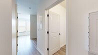 This single-floor unit offers a clean, contemporary design throughout, characterized by a bright and cohesive aesthetic. Light-toned hardwood-style flooring flows seamlessly across all areas of the space, creating a sense of continuity and warmth.

The hallway entrance connects the key areas of the unit efficiently, featuring panel-style interior doors with sleek matte black hardware — a modern contrast to the crisp white walls and trim. A small wall-mounted panel, likely an electrical or intercom fixture, is neatly integrated into the corridor.

The living area benefits from an impressive bank of large windows fitted with horizontal blinds, allowing for excellent natural light and outdoor views while maintaining privacy. The clean white walls and open layout give this room a spacious, airy feel.

The kitchen is well-appointed with a full suite of stainless steel appliances, including a refrigerator, electric range, over-the-range microwave, and dishwasher. Two-toned cabinetry — white upper cabinets and warm gray lower cabinets — provides generous storage, while a light countertop with an undermount sink completes the functional yet stylish design.

The bathroom maintains the same light and clean aesthetic, featuring warm wood-tone flooring, a glass-enclosed walk-in shower with a built-in bench, and classic white wall finishes. The overall ambiance of the space is modern, well-lit, and move-in ready.