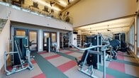 This fitness center offers a thoughtfully designed workout environment highlighted by soaring double-height ceilings and an open, airy atmosphere. The main floor is finished with a durable, colorful interlocking tile floor in teal, red, and beige tones, providing both visual interest and a resilient surface suited for high-traffic exercise areas. Large windows along one side of the space flood the interior with natural light while offering views of the surrounding greenery, creating an energizing workout environment. The main floor is outfitted with an extensive selection of Matrix-brand strength training machines, free weights, kettlebells, medicine balls, and a RAX functional training rack, along with dedicated storage cubbies for accessories. A separate cardio zone features spin bikes and additional aerobic equipment. A sleek glass-and-steel staircase with modern railings leads to a mezzanine level, which houses additional cardio machines such as ellipticals and steppers, all positioned beneath a ceiling fan for added airflow. The upper level overlooks the main training floor below through a glass balustrade. Dark-framed glass doors provide access to adjacent rooms, including what appears to be a meeting or lounge space. Wall-mounted mirrors and pendant lighting complete the functional yet polished aesthetic of this comprehensive fitness facility.