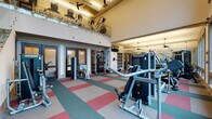 This fitness center offers a thoughtfully designed workout environment highlighted by soaring double-height ceilings and an open, airy atmosphere. The main floor is finished with a durable, colorful interlocking tile floor in teal, red, and beige tones, providing both visual interest and a resilient surface suited for high-traffic exercise areas. Large windows along one side of the space flood the interior with natural light while offering views of the surrounding greenery, creating an energizing workout environment. The main floor is outfitted with an extensive selection of Matrix-brand strength training machines, free weights, kettlebells, medicine balls, and a RAX functional training rack, along with dedicated storage cubbies for accessories. A separate cardio zone features spin bikes and additional aerobic equipment. A sleek glass-and-steel staircase with modern railings leads to a mezzanine level, which houses additional cardio machines such as ellipticals and steppers, all positioned beneath a ceiling fan for added airflow. The upper level overlooks the main training floor below through a glass balustrade. Dark-framed glass doors provide access to adjacent rooms, including what appears to be a meeting or lounge space. Wall-mounted mirrors and pendant lighting complete the functional yet polished aesthetic of this comprehensive fitness facility.