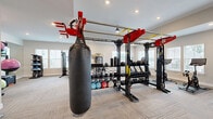 Located at 250 Battery Park Cir in Pendleton, SC, this fitness center offers a thoughtfully designed, single-floor workout space that accommodates a wide variety of training styles. The room is finished with neutral-toned carpet tiles in a herringbone pattern, recessed lighting, and multiple windows fitted with white blinds that allow ample natural light to fill the space.

The strength and functional training zone is anchored by a large multi-station rig in a bold red and black finish, supporting TRX suspension straps, a heavy punching bag, medicine balls, and an extensive dumbbell rack. A dedicated cable machine and weight bench provide additional options for resistance training. A wall-mounted mirror expands the sense of space and supports form-checking during workouts.

The cardio section features treadmills and stationary spin bikes, each positioned near the windows to provide an open, airy feel during use. Rowing machines and additional cardio equipment round out the options. A wall-mounted flat-screen TV is centrally positioned, enhancing the workout experience.

Stability balls, BOSU trainers, resistance tools, and balance equipment are neatly organized along the walls, ensuring the space remains uncluttered and functional. The clean, neutral aesthetic combined with a diverse equipment offering makes this fitness center a comprehensive facility for residents.