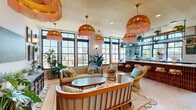 Located at 808 Constance Street in New Orleans' Lower Garden District, this single-floor commercial space offers a thoughtfully designed café and bar environment brimming with personality. The interior is defined by its retro-eclectic aesthetic, anchored by striking rattan and fringe pendant chandeliers in warm amber and blush pink tones that hang throughout the space, creating an inviting and playful atmosphere.

The dining area features terrazzo flooring, teal-and-white striped upholstered chairs paired with light wood frames, and marble-topped tables. A gallery wall of framed paintings and art pieces lines the white walls, complemented by lower decorative tilework and mirrored wall cutouts that add depth to the room. Lush potted plants are positioned throughout, adding a fresh, organic element.

The bar counter is clad in warm amber tiles with a white quartz countertop, and overhead glass-and-metal shelving displays glassware and plants. A comfortable lounge area features rattan and upholstered seating arranged around a central table, bathed in natural light pouring through large black-framed windows that offer elevated views of the surrounding New Orleans cityscape. The overall ambiance is vibrant, warm, and distinctly New Orleans in character.