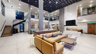 This contemporary two-story amenity space makes an immediate impression with its soaring double-height ceilings, exposed dark-painted structural beams, and large-format porcelain tile flooring throughout. The main level centers around a generous open lounge anchored by a striking three-sided glass fireplace set within a sleek marble-clad surround, complemented by a large wall-mounted TV above. Comfortable sectional seating in warm tan leather is arranged atop an abstract area rug, while sculptural linear light fixtures add artistic flair overhead. The open-plan layout seamlessly connects to a game and entertainment zone, complete with a foosball table, additional lounge seating, a large display screen, and a bar-height seating area set against a textured stone accent wall. A dedicated dining area with modern tables and chairs sits adjacent to a well-equipped kitchen, ideal for gathering and entertaining. Glass-partitioned rooms enclosed by gray brick facade walls house a private conference or meeting room with a poker table and accent seating. A secondary lounge offers a shuffleboard table, a TV, and direct access to an outdoor area through sliding glass doors. A floating staircase with wood treads and black metal railings connects to the upper mezzanine level, which overlooks the main floor below through decorative laser-cut screens.