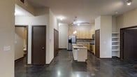 This single-floor space showcases a thoughtfully designed open-concept layout that seamlessly connects the kitchen and living areas. The kitchen serves as the focal point, featuring light wood cabinetry, a mosaic tile backsplash, and a full suite of stainless steel appliances, including a French door refrigerator with an ice and water dispenser, a built-in microwave, and a range. A center island with a sink and dark stone countertop provides both additional workspace and a natural gathering spot, illuminated by stylish pendant lighting overhead. Track lighting along the ceiling supplements the ambiance and ensures the space is well-lit throughout. The polished concrete floors extend across the entire unit, lending an industrial-modern aesthetic that is both durable and visually striking. Dark-stained interior doors create a bold contrast against the neutral walls, adding a layer of contemporary design. The open corridor leading into the space offers ample room for movement and transition, enhancing the overall sense of flow. This unit combines functional design with modern finishes, making it a highly practical and attractive living space.