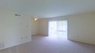 This two-floor space presents a clean, straightforward design centered around comfort and natural light. The main living area is finished with wall-to-wall carpeting in a neutral tone, contributing to a warm and quiet atmosphere. A ceiling fan is centrally positioned overhead, providing both lighting and air circulation throughout the room. One of the most notable features of this level is the large sliding glass door dressed with sheer vertical blinds, which floods the space with abundant natural light and provides direct access to an outdoor balcony. The balcony offers a glimpse of surrounding greenery, adding a pleasant outdoor connection to the interior. The walls are finished in a soft, neutral palette, maintaining a bright and airy feel throughout. The open layout of the main living area allows for flexible furniture arrangement and a sense of spaciousness. The overall design is straightforward and practical, with a neutral aesthetic that serves as a versatile backdrop. Spanning two floors, the space offers separation between living and private areas, making it well-suited for everyday functionality.