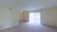 This two-floor space presents a clean, straightforward design centered around comfort and natural light. The main living area is finished with wall-to-wall carpeting in a neutral tone, contributing to a warm and quiet atmosphere. A ceiling fan is centrally positioned overhead, providing both lighting and air circulation throughout the room. One of the most notable features of this level is the large sliding glass door dressed with sheer vertical blinds, which floods the space with abundant natural light and provides direct access to an outdoor balcony. The balcony offers a glimpse of surrounding greenery, adding a pleasant outdoor connection to the interior. The walls are finished in a soft, neutral palette, maintaining a bright and airy feel throughout. The open layout of the main living area allows for flexible furniture arrangement and a sense of spaciousness. The overall design is straightforward and practical, with a neutral aesthetic that serves as a versatile backdrop. Spanning two floors, the space offers separation between living and private areas, making it well-suited for everyday functionality.