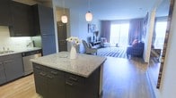 This well-appointed single-floor residence showcases a thoughtfully designed open-concept layout that seamlessly connects the kitchen and living areas. The kitchen serves as the heart of the space, anchored by a substantial island topped with light speckled granite countertops and complemented by dark espresso cabinetry with sleek hardware. Two elegant pendant lights hang above, casting a warm, inviting glow over the workspace. Full-height cabinetry provides generous storage, while the overall kitchen design balances both form and function. The open layout flows naturally into the living area, which features warm hardwood flooring throughout. The living space is bright and airy, benefiting from large floor-to-ceiling windows and sliding glass doors that lead to a private balcony, allowing an abundance of natural light to fill the interior. A bold geometric area rug defines the seating zone, adding visual interest and texture to the space. The neutral wall tones and soft drapery create a calm, cohesive aesthetic that ties the entire space together. The combination of contemporary finishes, practical design, and seamless indoor-outdoor connection makes this a comfortable and stylish living environment.