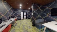Located at 90 New St in New Brunswick, NJ, this single-floor computer lab is a thoughtfully designed technology workspace that combines functional utility with striking contemporary aesthetics. The lab's most defining visual element is its bold, dark wall treatment featuring a vivid geometric pattern of intersecting lines rendered in white, red, and yellow — applied consistently throughout the space to create a cohesive, energetic atmosphere.

Warm wood slat ceilings run throughout the lab, providing an inviting contrast to the dark walls, while recessed lighting ensures the workspace remains well-illuminated. The floors are covered in a multi-toned mosaic carpet in shades of green, grey, and yellow, adding further visual dynamism underfoot.

The main area houses a row of desktop computer workstations positioned along a shared counter, accompanied by vibrant red ergonomic chairs. A dedicated printing station is equipped with an HP printer and an additional desktop terminal. A private meeting room, accessible through a glass-panel door, offers a compact table, seating, and a wall-mounted display screen — suitable for small group collaboration. Additional seating, including a grey sofa, provides a more relaxed area within the lab. Posted computer lab rules are visible near the entry, reinforcing the space's structured, communal purpose.