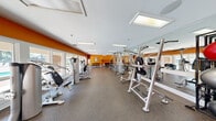 Located at 975 Westmeade Dr in Chesterfield, Missouri, this recreational facility offers a comprehensive range of amenities designed to support an active lifestyle. The generously sized main floor houses several distinct functional areas that flow cohesively throughout the space.

The fitness center is the centerpiece of the facility, characterized by bold orange accent walls, large windows that invite ample natural light, and floor-to-ceiling mirrors that enhance the sense of openness. The rubber-floored workout area is outfitted with an extensive selection of strength-training machines, cable stations, free weights with a full dumbbell rack, and cardiovascular equipment including ellipticals and steppers. Vending machines are conveniently located near the fitness zone for added accessibility.

Adjacent to the fitness center, a dedicated racquetball court features classic hardwood flooring with regulation court markings, bright overhead lighting, and clean white walls, providing an ideal environment for competitive play.

A welcoming entry area with tile flooring, decorative columns, and recessed lighting serves as a transitional hub connecting the fitness spaces to the outdoors. Large glass doors open directly to an expansive outdoor pool deck, where a freeform pool is surrounded by extensive lounge seating. The brick exterior architecture frames the outdoor space, lending a classic, polished character to the overall facility.