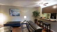 This single-floor residence offers a welcoming and functional layout designed for comfortable everyday living. The open-concept living area is anchored by rich hardwood-style flooring that extends seamlessly throughout the space, creating a warm and cohesive aesthetic. The living room provides ample room for relaxed seating, highlighted by a tufted leather sectional sofa and accent chair, complemented by a decorative floor lamp and a vibrant landscape painting that adds a pop of color to the neutral walls. A ceiling fan overhead ensures comfortable airflow throughout the space. A tall decorative plant adds a touch of greenery and natural charm to the corner of the room. Transitioning from the living area, a raised breakfast bar with dark wood cabinetry and bar stool seating defines the dining space, offering a casual spot for meals or conversation. The kitchen cabinetry features a dark espresso finish, providing a stylish contrast against the lighter walls. The textured ceiling adds a classic architectural detail throughout. Overall, the space strikes a balance between modern touches and cozy, livable design, making it an inviting and practical single-floor home.