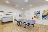 Located at 4659 Dawson Blvd in Atlanta, Georgia, this single-floor amenity space offers a well-appointed game room and a dedicated laundry facility. The game room serves as the centerpiece, featuring light wood-look flooring, crisp white walls, and recessed lighting that creates a clean, modern atmosphere. A regulation-size JOOLA ping-pong table anchors the room, providing ample space for gameplay. Colorful contemporary artwork adorns the walls, adding vibrancy and personality to the space. Large glass-paneled double doors along one wall flood the room with natural light and offer views of an outdoor pool area, seamlessly connecting interior and exterior amenities. A small seating nook with bright yellow chairs provides a comfortable spot for spectators or casual use. The game room also connects to a fitness area visible through the glass doors. The adjacent laundry facility is clean and efficiently designed, featuring a row of top-loading washers set on wood-look tile flooring beneath soft overhead lighting. A wide white counter surface runs alongside the machines, offering convenient space for sorting and folding. Together, these two amenity areas deliver a practical and engaging community environment suited for everyday living.