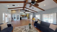This single-floor leasing office makes a strong first impression with its dramatic vaulted ceilings adorned with rich, exposed wood beams and multiple ceiling fans, creating an airy and comfortable atmosphere throughout. The main lounge area is generously proportioned, finished with warm hardwood-style flooring and painted in a cool, neutral gray palette that lends the space a calm, professional character.

A standout architectural feature is the floor-to-ceiling white brick fireplace, which anchors the lounge and adds a distinctive, residential charm to the office environment. Comfortable navy blue sofas, glass coffee tables, and patterned area rugs furnish the seating area, making it an inviting space for prospective residents. Large windows dressed with patterned curtains allow ample natural light to filter in throughout the day.

A dedicated workspace area with a desk and computer provides a functional zone for office tasks, while an information display stand keeps resident resources readily accessible. Toward the rear of the space, a full kitchen features warm wood cabinetry, a range, countertop appliances, and a white brick breakfast bar with bar stool seating — ideal for staff use or community gatherings. French doors on multiple sides of the space offer easy access and enhance the open, welcoming flow of the entire leasing office.