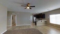 This single-floor residence offers an open-concept layout that effortlessly blends the living and kitchen areas into one cohesive, functional space. The living area features light hardwood-style flooring and neutral taupe walls, creating a warm and inviting atmosphere. A ceiling fan with an integrated light fixture serves as a practical centerpiece overhead, enhancing airflow and comfort throughout the space. The kitchen is a standout feature, showcasing rich dark cabinetry that contrasts elegantly with the light countertops and a clean white breakfast bar with seating overhang — ideal for casual dining or entertaining. Stainless steel appliances, including a refrigerator and microwave, add a modern, polished touch to the culinary space. Recessed lighting in the kitchen ensures the area is well-lit and functional. A transition from hardwood-style flooring to carpet in the living area adds textural variety and a cozy feel underfoot. The open floor plan allows for flexible furniture arrangements, maximizing the sense of space and flow. Doorways visible from the main living area suggest additional rooms, contributing to the overall practicality and livability of this well-designed single-level home.