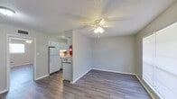 This single-floor unit offers a clean, functional layout with a cohesive neutral color palette and modern laminate wood flooring running throughout. The living area is open and well-proportioned, featuring a ceiling fan with light fixture, a textured ceiling, and a front entry door flanked by a large window that allows natural light to fill the space. The layout connects the living area directly to the galley-style kitchen, which is partially partitioned by a half-wall. The kitchen is equipped with white cabinetry, gray laminate countertops, a double-basin stainless steel sink, a refrigerator, and a range. A fire extinguisher is mounted at the kitchen entrance. The bedroom is a comfortable, well-sized room with its own ceiling fan, a window providing natural light, and a closet for storage. The laminate flooring continues seamlessly into the bedroom, maintaining a consistent feel throughout. The bathroom features a full tub and shower surround with clean white finishes and chrome fixtures. Clean white baseboards and neutral gray walls unify the space, giving the unit a bright, move-in-ready appearance that is both practical and inviting.