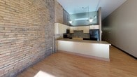 This single-floor unit showcases a compelling blend of industrial character and contemporary design. A defining feature throughout the space is the prominent exposed brick wall, which runs the length of the unit and lends warmth, texture, and historic charm to the overall aesthetic. Light hardwood flooring flows continuously across the main living areas, complementing the earthy tones of the brick and creating a cohesive, inviting atmosphere.

The main living area is open and well-proportioned, benefiting from natural light that streams through two windows fitted with roller shades. Track lighting overhead provides supplemental illumination, while the clean, neutral wall tones keep the space feeling bright and versatile.

The kitchen is a standout feature, designed in an L-shaped configuration with ample cabinetry in a light wood finish and dark laminate countertops. Black appliances, including a refrigerator, range, and microwave, are neatly integrated. Pendant lighting hangs above the kitchen area, adding an industrial-modern touch. The kitchen opens to the main living space via a peninsula counter, encouraging an open and connected layout.

An additional room, also accented by exposed brick and visible ductwork, provides flexible use as a bedroom or den. The bathroom is cleanly finished with tile flooring, a sliding glass shower enclosure, and modern fixtures.
