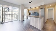 This well-appointed 1-bedroom, 1-bathroom unit offers a thoughtfully designed layout spread across a single floor. The open-concept kitchen and living area serves as the heart of the home, featuring warm wood-toned cabinetry, a sleek kitchen island with a stone countertop, stainless steel appliances, and pendant lighting. Floor-to-ceiling sliding glass doors flood the living space with abundant natural light while offering expansive urban views of the surrounding cityscape. Consistent hardwood-style flooring in a neutral grey tone flows seamlessly throughout the entire unit, lending a cohesive and modern aesthetic. A dedicated hallway connects the living areas to the private bedroom, which includes a built-in closet with integrated shelving, providing ample storage. The bedroom also benefits from large sliding glass doors with city views, creating a bright and airy atmosphere. The full bathroom is finished with large-format grey tile, a soaking tub with an overhead shower system, and contemporary wood-grain cabinetry with chrome fixtures. Clean white walls, recessed lighting, and crisp baseboards throughout the unit contribute to a polished, modern feel.