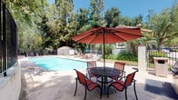 Located in the desirable community of Oak Park, California, this outdoor amenity space at 461 Savona Way offers a well-appointed recreational area designed for relaxation and leisure. The centerpiece is a generously sized swimming pool with crystal-clear blue water, complemented by an adjacent spa, providing residents with a resort-like experience. The pool deck is finished with durable concrete and features multiple red lounge chairs positioned to capture the abundant Southern California sunshine.

A shaded outdoor dining area with a round patio table, matching chairs, and a large red market umbrella provides a comfortable spot to gather and unwind. Nearby, a dedicated BBQ station offers a convenient space for outdoor grilling. The entire pool area is enclosed by a secure black iron fence, ensuring privacy and safety, with stucco pillar accents that echo the Spanish-influenced architectural style of the surrounding buildings, which feature characteristic red clay tile roofs.

The space is framed by tall, mature trees that provide natural shade and a lush, green backdrop, enhancing the tranquil ambiance. Well-maintained landscaping with a variety of shrubs, flowering plants, and potted greenery adds color and vitality throughout. A utility shed is neatly tucked within the fenced area. The overall atmosphere is serene, inviting, and ideally suited for enjoying the warm Oak Park climate year-round.