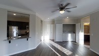 This single-story space showcases a thoughtfully designed open-concept layout that maximizes both functionality and flow. Dark hardwood floors run throughout, contrasting cleanly with the crisp white walls and trim. Crown molding adds a refined architectural detail along the ceiling line, while recessed lighting and a ceiling fan with an integrated light fixture ensure the space is well-lit and climate-comfortable.

The main living area is bright and open, with natural light streaming in and illuminating the dark floors. A dedicated laundry area is built into an alcove along one wall, housing a side-by-side washer and dryer unit — a convenient in-unit feature. Multiple interior doors provide access to additional rooms, suggesting a practical and versatile floor plan.

Adjacent to the living space, the kitchen is partially visible and features dark cabinetry, granite or stone countertops with a breakfast bar overhang, stainless steel appliances including a microwave, and a stovetop — offering a functional cooking environment that connects naturally to the living area. The overall ambiance is clean, modern, and move-in ready.
