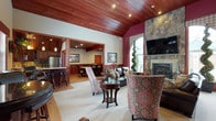 This single-floor clubhouse exudes warmth and sophistication through its thoughtfully designed interior. The main gathering area draws immediate attention with its vaulted ceiling clad in rich, stained wood planks, complemented by recessed lighting that adds a cozy ambiance. A striking floor-to-ceiling stone fireplace anchors one wall, topped by a mounted television and flanked by an arched window that invites natural light into the space. Elegant, patterned armchairs and leather seating are arranged around a classic wooden coffee table, creating an intimate and welcoming lounge area.

The space flows seamlessly into a formal dining area, visible through a wide open passage framed by warm wood millwork and wainscoting. This connected area features a dining table surrounded by chairs, ideal for group gatherings. Adjacent to the dining space is a well-appointed kitchen with bar-height seating, stainless steel appliances, and a pendant-lit counter, offering full hospitality functionality. Rich wood tones, plaid drapery, and carefully chosen décor throughout unify the space with a refined, lodge-inspired aesthetic.