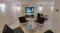 This single-floor space showcases a refined, contemporary design with a clean and sophisticated aesthetic. The centerpiece of the main lounge area is a linear gas fireplace set within a striking stone tile surround, above which a large wall-mounted flat-screen television is seamlessly integrated — creating a dual-purpose focal point that is both functional and visually impactful. Flanking the fireplace, two modern wall sconces provide warm, ambient lighting that complements the neutral tones of the space. The seating arrangement features a pair of classic Barcelona-style chairs in rich dark leather with polished chrome frames, paired with a round glass-topped coffee table — a combination that speaks to modernist design sensibilities. A matching ottoman anchors the foreground, adding to the lounge's comfort and cohesion. The flooring throughout consists of large-format light-colored tiles that enhance the sense of spaciousness and contribute to the room's bright, airy feel. Subtle wall tones in soft beige and white unify the space, while a small refreshment station is visible to the side, adding a practical touch. Overall, the space exudes a polished, upscale ambiance suited for both relaxation and social gatherings.
