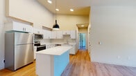 Located at 3546 Landview Dr SE in Olympia, WA, this well-designed 2-bedroom, 2-bathroom single-floor residence offers a clean, modern aesthetic throughout. The open-concept kitchen and living area serves as the heart of the home, boasting soaring ceilings, warm luxury vinyl plank flooring, and recessed lighting. The kitchen is outfitted with crisp white cabinetry featuring matte black hardware, white quartz countertops, a stainless steel sink with a gooseneck faucet, and a full suite of stainless steel appliances including a refrigerator, range, and microwave. A center island with pendant lighting provides additional prep space and casual dining options. A hallway connects the living area to the two bedrooms and bathrooms. The spacious primary bedroom features elevated ceilings, a ceiling fan, and access to a closet and adjoining bathroom. The second bedroom offers a ceiling fan, a wall-mounted heater, and a window with views of mature surrounding trees. Both bathrooms are finished with white vanities, tub-shower combinations, and light wood-tone flooring that flows consistently throughout the entire residence, creating a cohesive and inviting atmosphere.