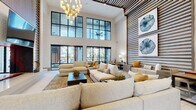 This sophisticated amenity space is defined by its dramatic double-height ceilings and an open, airy layout that creates a strong sense of grandeur throughout. The centerpiece of the space is a generously sized lounge area furnished with oversized, neutral-toned sectional sofas and accented with warm gold throw pillows, all anchored by a dark wood coffee table atop a textured area rug. Floor-to-ceiling steel-framed windows and glass doors flood the interior with natural light and offer views of an outdoor terrace, where lush palm trees and a covered patio with seating are visible. A statement chandelier with globe-shaped amber glass fixtures adds an elegant focal point above the main seating area. A distinctive horizontal wood slat accent wall runs throughout multiple areas, providing visual continuity and a warm, organic texture that contrasts beautifully with the crisp white walls. The game room features a full-size billiards table, a wall-mounted flat-screen television, and lounge seating, all beneath similarly soaring ceilings adorned with curated artwork. An adjacent work lounge area offers a functional desk and seating nook with direct access to the outdoor terrace through floor-to-ceiling glass doors. The kitchenette is outfitted with a long marble-topped island with bar seating, rich wood cabinetry, and a beverage station, completing this well-appointed communal environment.