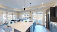 This well-appointed 2-bedroom, 2-bathroom residence offers a thoughtfully designed single-floor layout with modern finishes and a bright, open atmosphere. The combined kitchen and living area serves as the heart of the home, anchored by a large central island with a white quartz countertop and seating for multiple guests. The kitchen is outfitted with dark navy cabinetry accented by gold hardware, stainless steel appliances, a subway-tile backsplash, and ample storage. Pendant lighting and recessed fixtures illuminate the space, while oversized windows and access to an outdoor balcony flood the living area with natural light. A tray ceiling in the living room adds architectural dimension and depth. The primary bedroom is a serene retreat featuring plush carpeting, soft neutral tones, and generous proportions. The en-suite bathroom includes a dual-sink vanity with dark cabinetry, gold fixtures, and wood-look luxury vinyl flooring. The second bedroom is equally inviting, with large windows offering outdoor views and access to a dedicated closet. A second full bathroom features a tub-shower combination. Wood-look flooring runs through the main living areas, and Craftsman-style interior doors with black hardware provide a cohesive design detail throughout the residence.