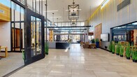 This contemporary amenity center is anchored by a striking, double-height lobby entrance defined by expansive floor-to-ceiling glass walls, polished large-format tile flooring, and a dramatic arrangement of geometric pendant light fixtures suspended from above. The open, airy atmosphere draws in abundant natural light, creating a bright and welcoming arrival experience.

Adjacent to the lobby, a stylishly appointed lounge area features a comfortable sectional sofa, backlit display shelving with curated décor, and sweeping views of the outdoor pool courtyard through oversized sliding glass doors. A nearby bar and social lounge offers bar-height seating beneath a warm wood-paneled ceiling, complete with a wall-mounted television and a dedicated beverage station with wine storage.

The fitness facilities are extensive and well-equipped. A spacious main gym features yellow-accented strength and cable machines, free weights, cardio equipment, and large windows overlooking the exterior. A separate training studio provides spin bikes, medicine balls, TRX equipment, and hardwood flooring ideal for functional workouts.

The leasing office area includes an open reception workspace, private consultation offices with glazed partitions, and comfortable seating with outdoor garden views. Throughout, the design language is consistently modern, combining warm wood tones, bold industrial metal framing, and bright, open sightlines.