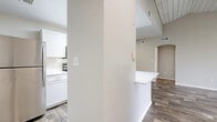 This single-story residence offers a thoughtfully designed layout encompassing a living room, kitchen, den, bedroom, bathroom, walk-in closet, and two hallways. The living room is a standout space, featuring a striking vaulted ceiling with wood-plank detailing and rich wood-look flooring throughout. A sliding glass door fills the room with natural light and provides outdoor access. The kitchen is outfitted with crisp white cabinetry, subway tile backsplash, white countertops, and stainless steel appliances including a refrigerator and range. A breakfast bar with a white countertop connects the kitchen to the adjacent dining or living area. The bedroom is carpeted and finished with crown molding and a ceiling fan, with a window that invites natural light and outdoor views. It connects to a generously sized walk-in closet equipped with wire shelving. The den offers additional flexible living space, also carpeted with crown molding. The bathroom features wood-look tile flooring, white cabinetry with a vanity mirror, and a tiled shower-tub combination. Neutral tones, arched doorways, and consistent finishes throughout lend the space a cohesive and polished character.