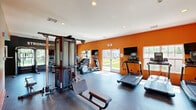 Located at 2800 New Brunswick Ave in Piscataway, NJ, this well-equipped fitness center offers a single-floor layout designed to support a comprehensive workout experience. The space is immediately distinguished by its bold, energizing orange accent walls and dark rubber flooring, which provide both visual impact and a functional, durable surface for exercise. Recessed lighting throughout keeps the room bright and well-illuminated.

The exercise room is outfitted with a variety of cardio equipment, including multiple treadmills and an elliptical machine, each positioned near large windows and glass doors that flood the space with natural light and offer views of the surrounding outdoor greenery and pool area. A multi-station cable machine and adjustable weight bench anchor the strength training zone, providing users with versatile options for resistance workouts. Wall-mounted flat-screen televisions are strategically placed for viewing during cardio sessions.

Motivational wall art, including the bold phrase "STRONGER THAN YESTERDAY," adds an inspiring atmosphere to the room. A seating bench near the entrance provides a practical resting area. The fitness center is accessed via glass doors and is clearly marked with posted fitness center rules, ensuring a safe and organized environment for all users.
