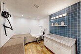Located at 2010 W Jackson St in Morton, Illinois, this thoughtfully designed dog grooming room delivers a fully equipped, professional-grade experience in a compact, single-floor layout. The space is accessible via a connecting hallway and opens into a well-lit grooming area finished with warm wood-look flooring and clean white walls.

The centerpiece of the room is a dedicated dog wash station featuring a low-entry tub with a handheld showerhead and an adjustable grooming stool, designed for ease of use with dogs of all sizes. A built-in diamond-plate metal ramp with a carpeted surface leads dogs gently up to the tub level, eliminating the need for lifting. A wall-mounted pet dryer is positioned nearby for a seamless post-bath routine.

Adjacent to the wash station, a bold blue ceramic tile accent wall provides a striking visual focal point. Set against it is a white cabinetry unit topped with a granite countertop, offering ample prep and storage space. An open floating shelf above displays grooming supplies and decorative accents, illuminated by a horizontal vanity light bar.

The room is completed by recessed ceiling lighting, a textured ceiling, and a whimsical wall quote that reinforces the space's dedicated canine purpose. A hallway connects the grooming room to the broader facility, making for a smooth and functional flow.